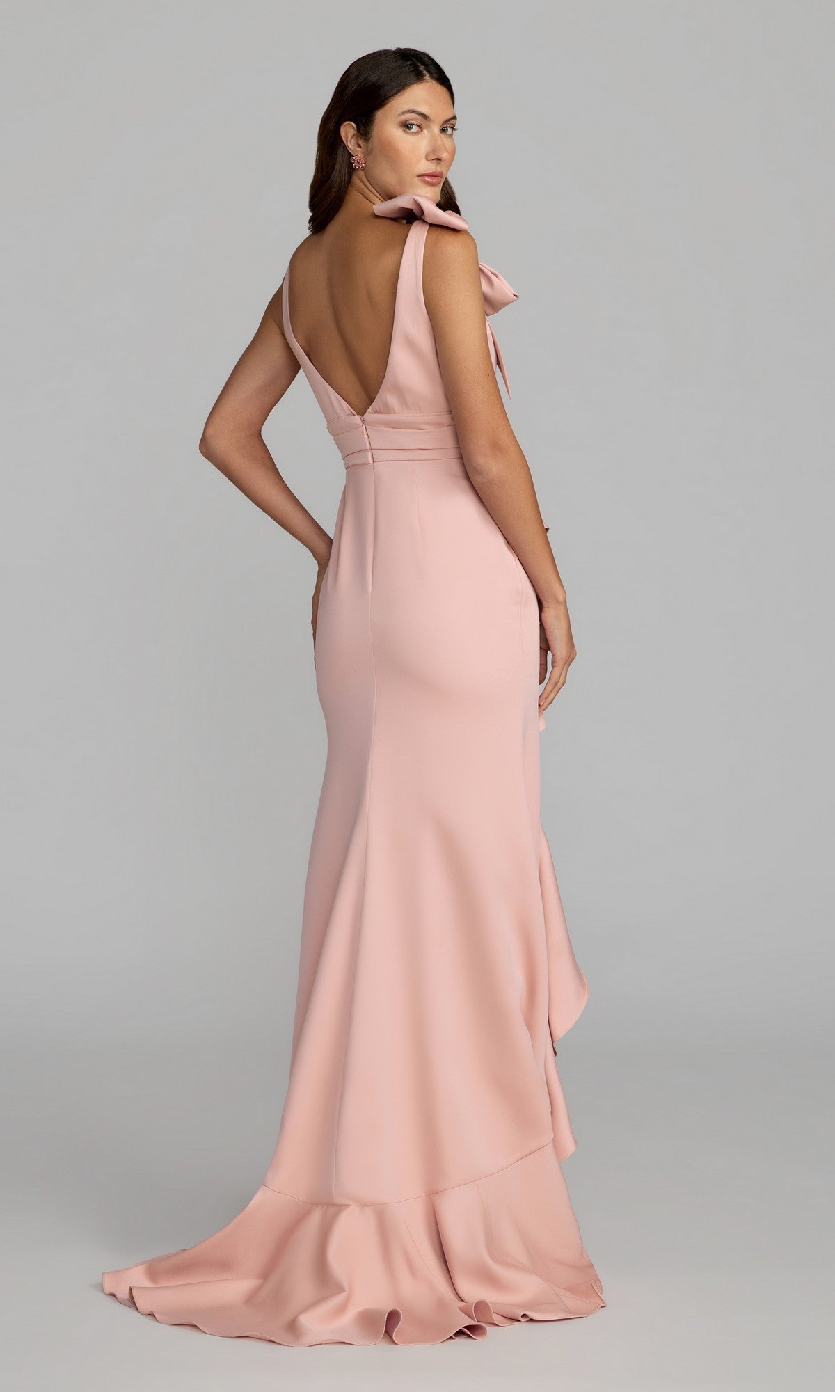 A woman stands against a plain gray background wearing the Mac Duggal 49965, a floor-length formal dress in blush pink featuring a deep V-back and ruffle detail.